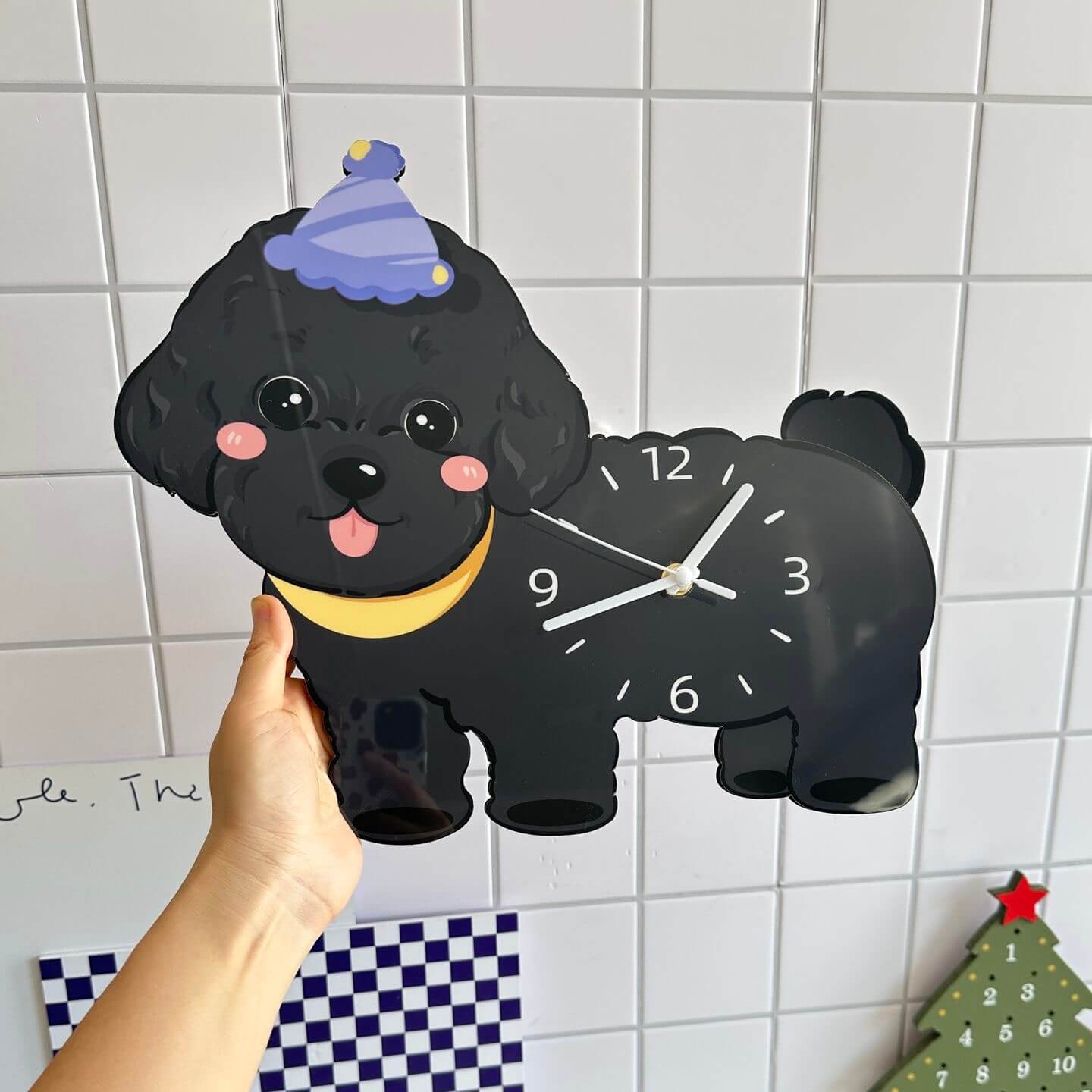 Black poodle dog clock held in hand on white tile wall with Christmas tree and checkered cloth